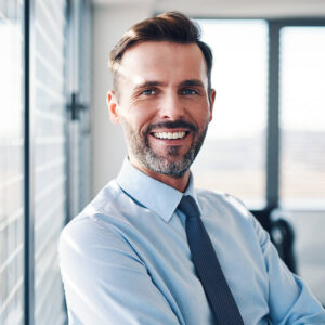 Mann in Business-Kleidung steht am Fenster und schaut lächeln in die Kamera