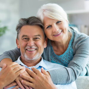 Ältere Frau umarmt älteren Mann von hinten, beide lächeln in die Kamera.